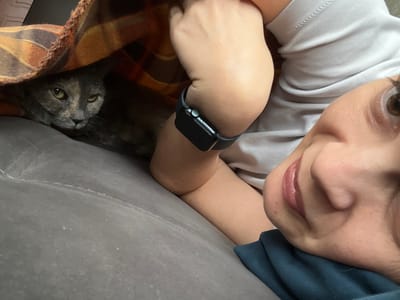 A close-up, low-angle shot shows a person and a cat peeking out from under an orange and brown plaid blanket on a gray couch. The cat, partially obscured by the blanket, has grey fur with lighter patches and green eyes. Its head is turned towards the camera, with an alert expression. The person's face is in the foreground on the right, with their eyes looking directly at the camera. Their mouth is slightly open, revealing teeth and tongue. They are wearing a light gray t-shirt and a dark blue garment around their neck. A black smartwatch is visible on their left wrist. The background consists of the soft texture of the couch and the plaid blanket. The image was taken indoors. The location is not specified in Tolyatti, Russia.