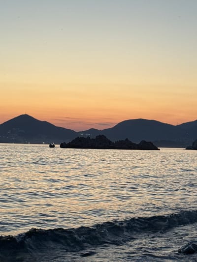 The image depicts a serene coastal scene at dusk, likely in Sveti Stefan, Montenegro. The sky transitions from a pale blue at the top to a warm gradient of orange and yellow near the horizon. Silhouetted mountains form the backdrop, with faint lights dotting the slopes of one, suggesting a town or settlement. In the foreground, the calm sea reflects the soft colors of the sky, with gentle waves lapping at a dark, rocky shoreline. A small, dark boat is visible in the middle ground, positioned on the water between the shore and a cluster of jagged rocks that emerge from the sea. The overall atmosphere is tranquil and picturesque.