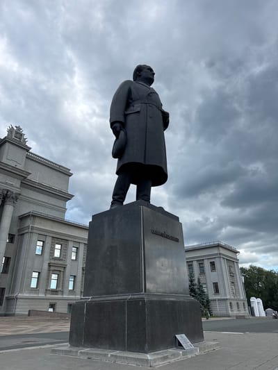 The image depicts a statue of a man in a long coat, holding a hat in his left hand. The statue stands on a tall, dark pedestal with an inscription in Cyrillic script. To the left of the statue is a large, gray building with classical architectural elements, including columns and ornate detailing. To the right, another gray building is visible, along with evergreen trees and a cloudy sky. The scene appears to be a public square or plaza, with paving stones in the foreground. The statue is located in Novokuybyshevsk, Russia.