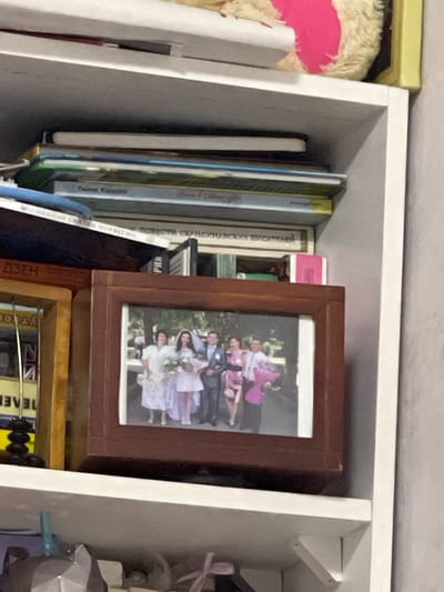 This image shows a bookshelf in a room. On the top shelf, there are stacks of books and a stuffed animal. The middle shelf holds a framed photograph of a wedding party, with a bride, groom, and other guests. To the left of the photograph, a yellow framed object is partially visible. The bottom shelf contains some decorative items. The image quality is poor, making it difficult to discern specific details about the objects or people. There is no indication of the city of Donskoye, Russia.