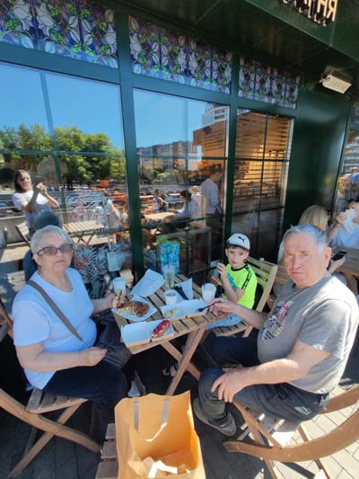 In this image, a group of people are enjoying a meal outdoors at a cafe. The scene is set on a sunny day, as evidenced by the bright light and clear blue sky reflected in the large windows of the establishment. The cafe appears to be in an urban setting, with buildings visible in the background reflections.

The foreground features three individuals seated at a small wooden table. On the left, an elderly woman with glasses and short gray hair is holding a piece of food and looking towards the camera. In the center, a young boy wearing a white cap and a bright green t-shirt is also looking towards the camera, with a fork in his hand. To the right, an elderly man with gray hair is seated, also looking at the camera.

The table is laden with food and drinks, suggesting a leisurely meal. There are pastries, drinks in cups, and packaging on the table. A large orange paper bag sits on the floor next to the table. The reflections in the window reveal more people and tables inside and outside the cafe, indicating it's a popular spot.

Visible text in the image includes Cyrillic script on the cafe's window, which appears to be a menu with items like "ЧАЙ ЧЕРНАЯ СМОРОДИНА" (Blackcurrant Tea) and "ЧАЙ ИМБИРНЫЙ С МЯТОЙ" (Ginger Mint Tea), suggesting the cafe may specialize in teas. The overall atmosphere seems relaxed and pleasant, with people enjoying good weather and food. The presence of both older and younger individuals suggests a multi-generational outing.