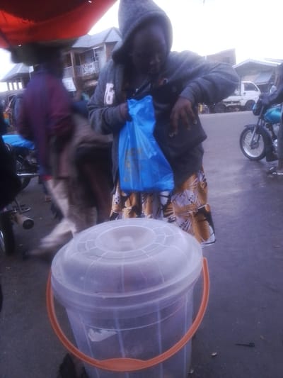 The media file shows a busy outdoor scene, likely a market in Gwong, Nigeria. In the foreground, a large, clear plastic bucket with an orange handle is prominently displayed. The bucket is partially filled with what appears to be goods. A person, seen from the waist down, is standing next to the bucket, holding a bright blue plastic bag. This individual is wearing a patterned skirt with geometric designs in yellow, white, and black.
Behind this individual, other people are blurred due to motion, suggesting a dynamic environment. One person is visible wearing a grey hooded jacket, with the hood up, and a dark vest over it. They are reaching into the blue plastic bag. In the background, there are buildings, a white truck, and a motorcycle, indicating a commercial area. The lighting suggests it is either early morning or late afternoon, with overcast skies. The overall impression is one of everyday commerce and activity. There is no discernible text in the image.