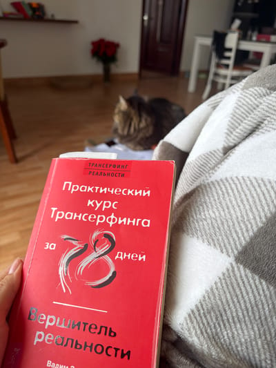 A person's hand holds an open red book with Russian text. The book's title appears to be "Практический курс Трансерфинга" which translates to "Practical Course of Transurfing". The book also features the number "78" prominently displayed in a stylized font. The person is wrapped in a grey and white checkered blanket. In the background, a cat is visible, and a bouquet of red roses in a vase is on a wooden floor. A shelf with books is mounted on the wall. The scene is indoors, in what appears to be a domestic setting. There are no visual cues to suggest this image was taken in Lâm Viên - Đà Lạt, Vietnam.