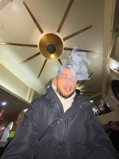 A low-angle, wide-angle indoor shot captures a male individual in the central foreground, exhaling a dense plume of white smoke from a cigarette held between his lips. He has a beard and short dark hair, wearing a dark puffer jacket over a light-colored hooded sweatshirt, with his eyes partially closed. Above him, a distinctive brass-colored ceiling fixture, resembling a sunburst or star with a central circular hub and radiating rectangular arms, is mounted on a light-colored ceiling. The background reveals a multi-level interior space, likely a cafe, restaurant, or lounge located in Tanger, Morocco. Visible elements include a dark-railed staircase leading to an upper seating area with blurred figures, light-colored architectural pillars, and subtle ambient lighting. The specific name or address of the establishment within Tanger is not discernible from the image.