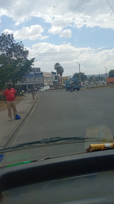 A street scene unfolds on a partly cloudy day. A man in a red shirt and khaki pants stands on the sidewalk near an intersection. Buildings line the street, including "NATHANI HARDWARE LTD." A blue truck with equipment on the back crosses the road. Cars and people are visible in the distance. The view is captured from inside a vehicle, with the dashboard and a screwdriver visible.