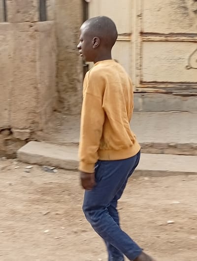 A single young male individual is depicted walking from right to left across an unpaved, dusty area in Gwagwarwa, Nigeria. The individual, appearing to be a boy with dark skin and short-cropped hair, is wearing a loose-fitting, long-sleeved mustard yellow sweatshirt and dark blue trousers. His head is turned slightly to his left, and his bare feet exhibit motion blur, consistent with movement. In the mid-ground, a low, rectangular concrete or stone slab rests on the ground. The background features a weathered, light brown concrete wall to the left and a large, light-colored metallic gate or door with visible structural framing to the right. The scene is illuminated by natural daylight.