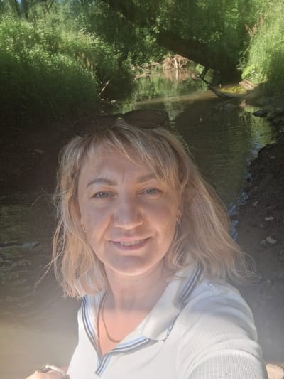 In the city of Дзяржынскі сельскі Савет, Belarus, a woman with blonde hair and sunglasses perched on her head takes a selfie in front of a creek. She is wearing a white collared shirt with blue and grey stripes around the collar. The creek flows through a wooded area with lush green trees and foliage lining its banks. The water is calm, reflecting the surrounding greenery. The sunlight creates a bright, hazy effect in the foreground of the image.