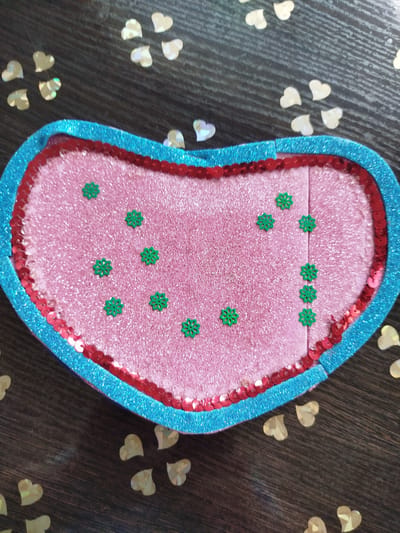 This is a close-up, top-down shot of a heart-shaped object against a dark wooden surface. The heart is outlined with a blue, glittery material, followed by a layer of red sequins. The interior of the heart is filled with a pink, glittery material and decorated with numerous small, green, star-shaped confetti. Scattered around the heart on the wooden surface are many small, golden heart-shaped confetti pieces. The scene is well-lit, highlighting the textures and colors of the heart-shaped object and confetti. The location is in Крывасельскі сельскі Савет, Belarus.