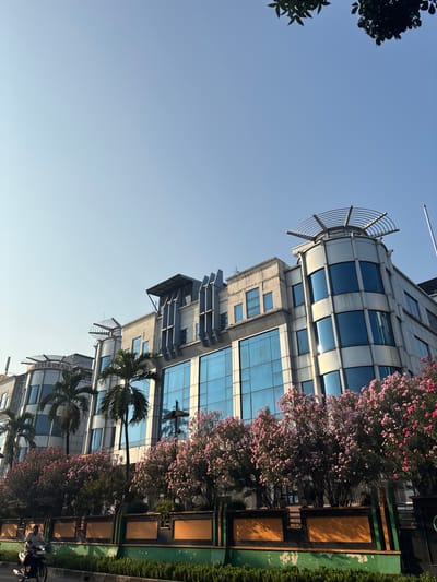 This is a low-angle, medium shot of a modern building in West Jakarta, Indonesia, under a clear blue sky. The building is a multi-story structure with a facade of blue glass windows and light-colored concrete or metal panels. The building has several architectural elements, including rounded corners and a circular structure with a metal frame on the rooftop of one section. Palm trees are visible in front of the building, and a line of blooming pink trees lines the foreground. A person is riding a motorcycle on a road in the lower left corner of the image, moving from left to right. A green median with decorative panels separates the road from the building and landscaping. The overall impression is one of an urban environment with commercial buildings and greenery.
