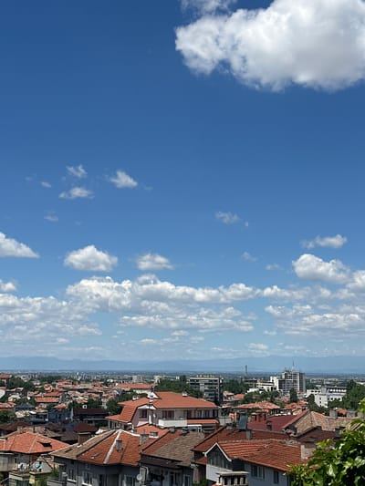 This is an eye-level, medium shot of a city in Asenovgrad, Bulgaria on a sunny day with a blue sky filled with white, puffy clouds. The foreground is dominated by the terracotta tiled roofs of numerous buildings, some of which are residential homes and others appear to be apartment buildings. The roofs have varying shades of red-brown and show signs of age. Interspersed with the rooftops are some trees and green foliage, particularly on the right side of the frame.
In the mid-ground, the cityscape continues with more buildings, some of which are taller, modern structures with white and grey facades, including some apartment blocks and office buildings. There are also some industrial-looking structures visible in the distance, such as a tall, slender tower. The far distance is marked by a range of hazy, blue mountains, suggesting a natural landscape surrounding the city. The overall impression is one of a densely populated urban area under a bright, cloud-filled sky. No people are visible in the image.