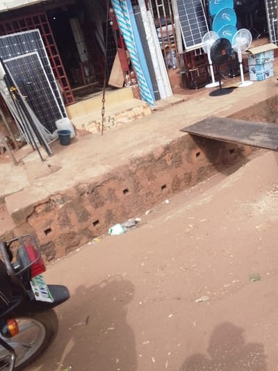 The image displays an outdoor commercial area in Ankpa, Nigeria. In the background, a storefront with a reddish-brown facade and a metal security gate exhibits various merchandise. Visible items include multiple solar panels, four pedestal fans (three white, one black), several blue circular satellite dishes, and bundles of striped blue, white, and grey pipes or conduits. A white jerrycan and a blue bucket are situated near the store's tiled entrance steps. A metal chain hangs vertically near the steps.
In the mid-ground, a reddish-brown concrete or brick drainage ditch runs parallel to the storefront, featuring several square openings along its wall. A dark metal plank bridges a section of this ditch. The foreground is an unpaved, reddish-brown dirt road surface, containing minor debris and sparse green vegetation. The rear section of a black motorcycle, displaying its taillight, fender, and part of the rear wheel with a partially visible license plate ("IM-134"), is present in the lower-left corner. Two distinct human shadows are cast upon the dirt road in the lower-right foreground.
