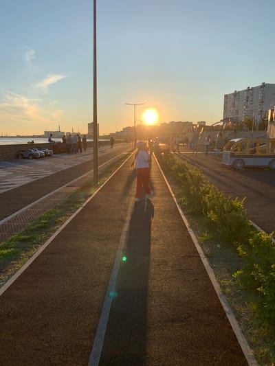A photograph taken in Nizhnevartovsk, Russia, captures a scenic outdoor scene at sunset. The image is oriented vertically, with the sun low on the horizon, casting a warm, golden glow across the landscape. The foreground features a paved pathway, likely a promenade or park trail, bordered by a strip of greenery and then a textured area with patterned paving stones.
Several people are visible along the pathway. In the middle ground, a person wearing orange pants and a white top is walking away from the camera, their shadow stretching long across the path. Further along, a group of people are gathered near what appears to be a playground or recreational area, with structures suggesting swings and climbing frames. Some of these individuals are standing, while others are seated. In the background, there are multi-story buildings, characteristic of an urban environment, bathed in the soft light of the setting sun.
The time of day is clearly sunset, indicated by the low position of the sun and the warm, elongated shadows. The sky is a clear blue with some wispy clouds towards the horizon. The presence of people engaged in leisurely activities, the paved pathways, and the park-like greenery suggest a public recreational space. The overall mood is peaceful and serene, enhanced by the beautiful sunset lighting. No visible text is present in the image.