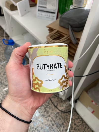 A person's hand, wearing a black beaded bracelet on the wrist, holds a cylindrical container of "BUTYRATE" food supplement. The container has a light green and brown abstract design with gold accents and text. The text on the container reads "BUTYRATE," "FOOD SUPPLEMENT," "600 mg SODIUM BUTYRATE," and "60 CAPSULES." The background suggests an indoor environment with shelves, stacked paper bags, a water bottle with a blue cap, and electrical cords. The image is taken from a close-up perspective, focusing on the hand and the product.