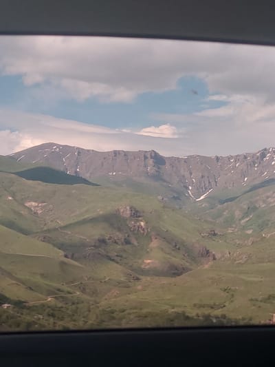 The image captures a panoramic view of a mountainous landscape, likely seen from a window, as indicated by the dark vertical frames on the left and right edges. The orientation of the image is vertical, presenting the sky and clouds on the left side and the extensive mountain range and green slopes on the right.
The left side of the frame features a bright, partly cloudy sky, with patches of light blue intermingled with soft white and light grey clouds. A small, dark speck, possibly a bug or debris on the window surface, is noticeable against the sky in the upper left quadrant.
Dominating the central and right portions of the image is a rugged mountain range. The mountains display a palette of dark grey and brown rocky surfaces, characteristic of exposed rock. Scattered patches of white, indicative of snow, are visible along the higher ridges and in various depressions of the peaks, suggesting a significant elevation or a recent snowfall. Below the rocky peaks, the lower slopes of the mountains are blanketed in vibrant green vegetation, appearing as lush grasslands or low-lying foliage. The landscape unfolds with rolling hills and valleys covered in green, extending towards the foreground on the bottom right.
The scene is brightly lit, indicating it is daytime with clear visibility. No specific activity or event is depicted, rather it is a static observation of natural scenery. There is no visible text within the image. Given the context of Yelpin, Armenia, this mountainous terrain is consistent with the region's geography.