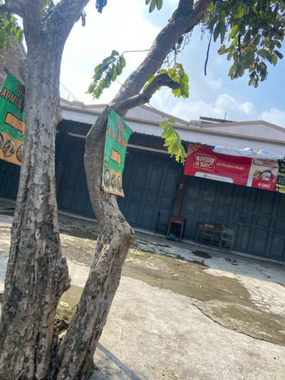 The image shows a tree trunk in the foreground, with a concrete driveway and a building in the background. The tree trunk is large and brown, with rough bark. It is split into two main branches. The driveway is cracked and gray, with a few patches of green moss. The building is a single story, with gray siding and several windows. There are two signs on the building, one for a store called "Toko SRC Al Mubarakah," and another for a store called "Sumber Berkah Mart." The signs are both red and white. There is no one present in the image. The location is a residential area in the city of Margaasih, Indonesia, with coordinates -6.942 latitude and 107.551 longitude.