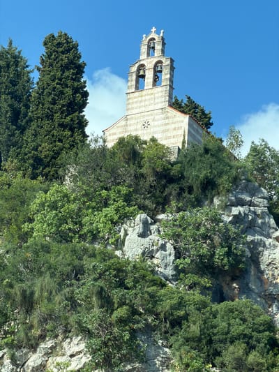 An image, correctly oriented, captures a stone church nestled on a rugged, tree-covered hillside under a bright blue sky with scattered white clouds. The setting is Kaluđerac, Montenegro, known for its scenic coastal and mountainous landscapes.
The central subject is a traditional church, constructed from light-colored stone blocks. Its most prominent feature is a tall, ornate bell tower (campanile) rising from its left side, composed of three visible levels of arched openings, each housing one or more dark bells. The church roof is terracotta-tiled. A circular decorative element, possibly a rosette window, is visible on the main facade of the church building.
The church is surrounded by dense, varied green vegetation. Taller, darker coniferous trees, likely cypresses, are visible on the lower left foreground, while a mix of lighter green bushes and smaller trees cling to the rocky terrain. Jagged, gray rock formations and cliffs form the backdrop and integrate with the vegetation around the church, suggesting it is built directly into the natural slope.
The time of day appears to be bright daytime, likely late morning or early afternoon, with clear weather and ample sunlight casting subtle shadows. There is no human activity or visible text in the image. The scene conveys a sense of tranquility and the harmonious integration of ancient architecture within a wild, natural environment.