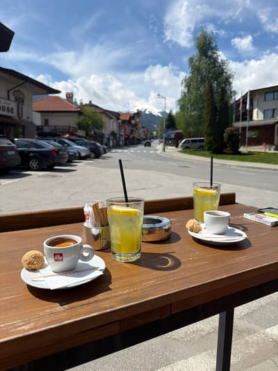 The image depicts an outdoor café scene in Bansko, Bulgaria. Two sets of drinks are arranged on a wooden table: each set consists of a small white cup of espresso on a saucer, accompanied by a small round biscuit, and a tall glass of yellow beverage with a black straw, possibly lemonade. An ashtray and condiment packets are also on the table. The background shows a street scene, with parked cars lining the side of the road, buildings with red-tiled roofs, and greenery. The sky is bright blue with scattered clouds.