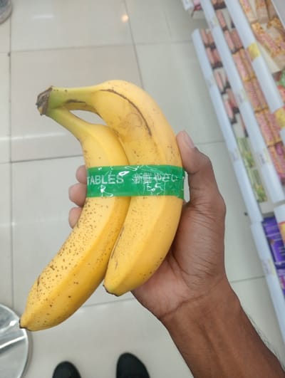 A hand is holding two ripe bananas, which are banded together with a green plastic wrap. The wrap has white text that includes "TABLES" and some Japanese characters. The bananas are yellow with some brown spots, indicating ripeness. The background shows a tiled floor and a retail shelf with various packaged goods, suggesting a grocery store or supermarket setting. The lighting appears to be artificial, typical of an indoor retail environment. The overall scene suggests a casual moment of selecting produce.