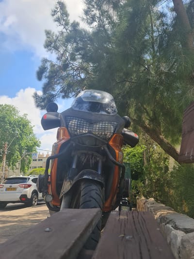A low-angle, close-up, full shot shows the front of an orange and black motorcycle parked next to a wooden bench. The motorcycle has a large windshield and prominent headlights. Behind the motorcycle, a white Subaru Crosstrek SUV is parked on the street. A pedestrian crossing sign is visible in the background, along with buildings and trees. The scene appears to be outdoors on a sunny day, with a blue sky and white clouds overhead. The overall impression is of a quiet street scene in a residential area. This image was captured in Haifa, Israel.