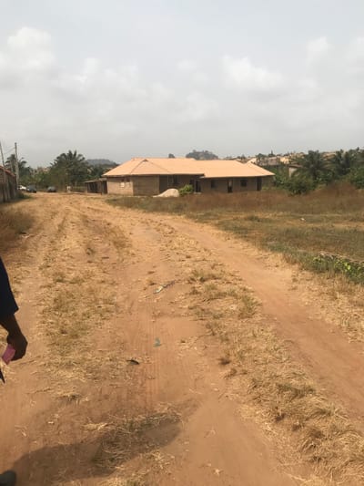 The image depicts an unpaved, reddish-brown dirt road in Akure, Nigeria, under a uniform overcast sky. The road exhibits distinct tire tracks and is interspersed with patches of dry, light-brown grass and sparse green vegetation. Scattered debris, including small pieces of plastic, is visible on the road surface. In the midground, a prominent single-story building constructed from grey concrete blocks features a distinct orange-brown corrugated metal roof. This structure appears to be under construction, with open window frames and no glass installed, and a visible pile of sand or aggregate positioned near its front. To the left of the main building, further down the road, several smaller structures and two parked vehicles are discernible. The background features undulating terrain with hills covered in dense green vegetation and additional distant buildings. In the immediate foreground, partially visible in the bottom left, is the lower right arm and hand of an individual wearing a dark blue sleeve, holding a small, rectangular pink object. No direct action or interaction is depicted by the person.