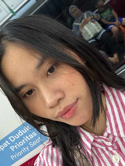 A close-up, selfie-style, head-and-shoulders shot shows a young woman with long, dark hair and light skin. She is wearing a pink and white striped shirt. Her hair falls across her face and shoulders. To the left of her, a blue sign with white text reads "pat Duduk Prioritas Priority Seat". In the background, out of focus, are other passengers on what appears to be public transportation. One woman wears a light blue hijab and holds a light green and white tote bag. Another person, a male, is seated next to her. The environment suggests an indoor setting like a train or bus.