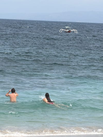 The image depicts a serene ocean scene with two individuals enjoying the water near the shore. The water transitions from a deep, dark blue to a lighter, turquoise hue as it approaches the beach, where gentle waves create a foamy white line along the sand.
In the distance, a white drone with a red accent is visible hovering over the water, suggesting a contemporary element to the scene. In the foreground, a person is submerged in the water up to their waist, facing away from the camera and looking towards the horizon. Their back is tanned and muscular, and they appear to be relaxed. Slightly further out in the water, another person, a woman with long dark hair, is swimming, also facing away from the camera. She is wearing a red bikini.
The overall atmosphere is peaceful and leisurely, with the warm tones of the sun illuminating the water and the skin of the swimmers. The time of day appears to be daytime, likely late morning or early afternoon, given the brightness of the light. The weather seems pleasant, with calm waters and clear skies suggested by the overall lighting. The location, Manggis, Indonesia, evokes a tropical beach setting. There is no visible text in the image.
