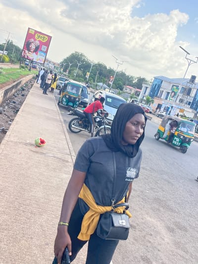 A young woman with dark hair and a black hijab is the main subject of the image, which appears to be taken outdoors in a busy urban environment, likely in Barakin Lerawi, Nigeria, given the context. She is wearing a dark grey t-shirt with a zebra-like graphic and the text "Quality Da" visible, and black pants. A yellow scarf is tied around her waist, and she has a black crossbody bag. She is looking towards the camera with a neutral expression.
The setting is a street with traffic. Several vehicles are visible, including a green and yellow tricycle (also known as an "okada" or "kekè") with license plate "PTA-10199", other tricycles, motorcycles, and cars. A billboard advertising "Yes! Cornflakes" is prominently displayed on the left side of the image, featuring a woman with curly hair holding a bowl of cereal. There are also buildings and trees in the background, suggesting a commercial or residential area. The sky is partly cloudy, indicating it might be daytime. The ground is paved, and there is some litter visible along the side of the road. A partially eaten piece of fruit, possibly a watermelon, lies on the pavement near the woman. The overall scene suggests a typical day in a bustling Nigerian town.