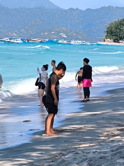 The image depicts a crowded beach scene in Indonesia. The water is a vibrant turquoise, with gentle waves lapping at the shore. In the background, a range of mountains forms a dramatic backdrop. Several boats are anchored offshore, suggesting the presence of tourist activities. On the beach, a group of people are visible, some wading in the shallows and others standing on the sand. The foreground is dominated by the sandy beach, with scattered shadows cast by palm trees. The overall impression is one of a lively and scenic beach destination.