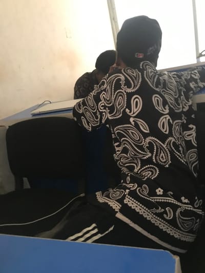 The scene is located in Calabar, Nigeria. A person is observed from behind, seated and leaning forward at a table or desk. The individual wears a black beanie, a long-sleeved black shirt with an intricate white paisley pattern, and black pants featuring two prominent white stripes on the outer leg, indicative of athletic wear. A silver or white laptop, closed, rests on a light blue table surface in front of the individual. Behind the seated person, a reflection of another individual or the same person is visible in a reflective surface on a cream-colored wall, showing a dark, possibly patterned garment. The background consists of a plain, light-colored wall with some visible discoloration or staining. The foreground includes a dark blue or black chair and a portion of a blue surface, likely part of the table.