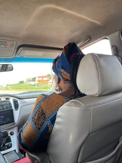 The image depicts a woman seated in the passenger seat of a car, facing away from the camera. She is wearing a colorful headwrap with blue and orange geometric patterns and a matching dress. Her hair is braided and hangs down her back. She is wearing gold earrings and a thin gold chain around her neck.

The interior of the car is visible, with light-colored leather seats and a beige headliner. The car's dashboard is partially visible, showing a screen with the number "70" displayed. The scene appears to be set in Sabojibi, Nigeria, given the context.

Outside the car, through the windshield, a bright, sunny day is indicated by the strong light. The background shows greenery, trees, and some buildings, suggesting a semi-urban or rural setting. The image is oriented sideways.