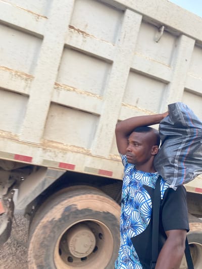 A man stands next to a large truck in Abofifia, Nigeria. The man is carrying a grey plastic bag on his head. He is wearing a blue and white patterned shirt, black shorts, and a black satchel. The truck appears to be a dump truck, with a white bed marked with square patterns and red reflective tape. The truck's tires are dusty. The ground appears to be dirt or gravel.