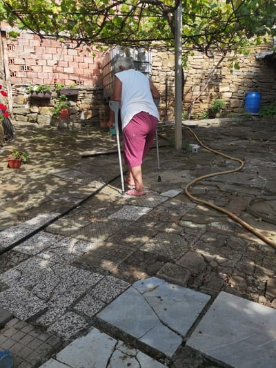 An elderly person with gray hair is walking with crutches in an outdoor courtyard in Lomets, Bulgaria. The person is wearing a white t-shirt and pink shorts, and pink sandals. The courtyard is paved with stone slabs, some of which are cracked. There is a hose lying on the ground and a large water tank in the background. The walls are made of stone and brick. Vines are growing on a trellis overhead, casting shadows on the ground.