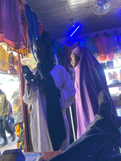 This image depicts an indoor market stall in Samaru, Nigeria, illuminated primarily by artificial blue light from an overhead LED strip. Two young females are prominent in the foreground. The female on the left, with dark skin, wears a black hijab, a white long-sleeved garment, and a black scarf. She is engrossed in a black smartphone held in both hands, with a red bracelet visible on her left wrist and a small light-colored bag hanging from her arm. The female on the right, also with dark skin, wears a light purple full-length hijab that covers her head and body; she is looking towards the right with an upward gaze, appearing to observe something beyond the frame.
The background features numerous colorful garments, including dresses and hijabs, displayed on racks and shelves, consistent with a clothing shop. A white ceiling fan with blue blades is mounted in the upper right. A dark wooden counter or surface is visible in the immediate bottom foreground, with the foot of a person wearing a dark sock and a decorative ring, suggesting the viewpoint of a seated individual. Through an open doorway or entrance on the far left, a male figure wearing a light green t-shirt, blue trousers, and sandals is partially visible, walking past. Another obscured figure in patterned clothing is behind him. The ceiling appears to be made of corrugated metal or a similar material.