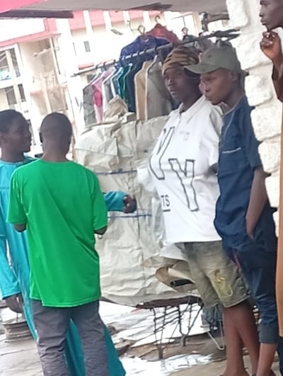 The image depicts a street market scene in Shaba, Nigeria, with a group of young men gathered. In the foreground, from left to right, a young man in a blue-green robe is looking to his left. Next to him, facing away from the camera, is another young man wearing a bright green t-shirt and dark gray pants. To their right, a young man in a white t-shirt with a graphic design of the letters "KY" in black outlined letters and the word "TS" below, is looking towards the right. He is wearing a patterned headwrap. Beside him, a young man in a dark blue shirt and a green baseball cap is also looking to his right. Partially visible on the far right is a person wearing a brown garment. Behind the individuals, clothing items are displayed on hangers, suggesting a clothing stall. The background shows buildings and a general street environment. The ground is covered with what appears to be debris and some metallic structures.