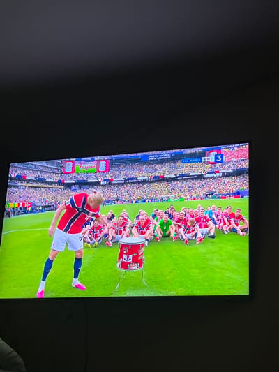 The image shows a television screen displaying a soccer match. A player in a red and blue jersey with the number 9 on his shorts is standing on a green field, bending over. Several other players in similar jerseys are sitting or kneeling behind him. To the right, a red drum with white and blue accents is placed on a stand. The background is filled with a stadium crowd and advertising boards. The scoreboard in the upper right corner shows "FIFA WORLD CUP QUALIFIERS" and "3". The image does not contain any elements that would indicate the location is the city of Dobrich, Bulgaria.
