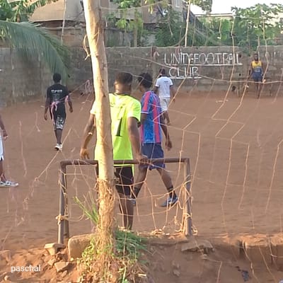 A dirt field in Nsugpe, Nigeria, shows several young men, possibly playing or practicing soccer. The backdrop displays a cement wall with "UNITY FOOTBALL CLUB" painted on it. One man wears a bright yellow jersey with the number "1" on the back. A soccer net stands in the foreground, partially obscuring the view. The lighting suggests it is late afternoon or early evening.