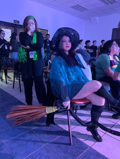 In the city of Arica, Chile, this image captures a scene that appears to be an indoor event. The primary focus is on two women dressed in costumes. One woman, in the foreground and center, is seated and wearing a large black witch's hat, a blue shawl with fringe, and black knee-high boots. She is holding a broom in her lap, with its bristles pointed towards the lower left of the frame. She is looking to her right, with her legs crossed. To her left, another woman is standing and appears to be crying or distressed, covering her mouth with her hand. She is wearing a green headband, a green ribbon around her neck, and black clothing. She has green pom-poms attached to her outfit. In the background, several other people are visible, standing and watching something off-camera. The environment appears to be a room with tiled flooring, a ceiling with fluorescent lights, and a sign on the wall that reads "SALIDA" (EXIT) with an arrow pointing left. The lighting in the room is predominantly blue.