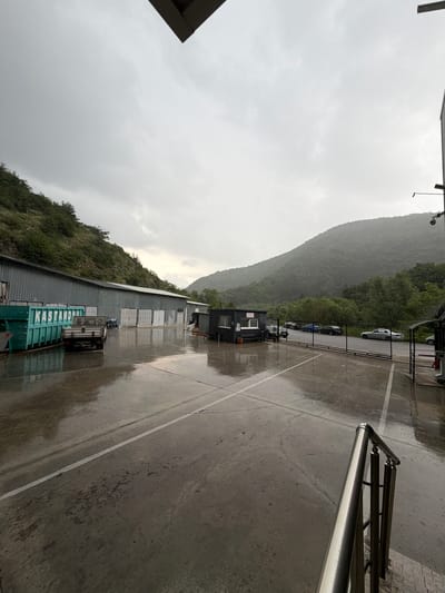 The image captures an outdoor scene dominated by natural landscape and industrial structures under an overcast sky. A large, steep, tree-covered mountain or hill occupies the left side of the frame, its verdant slopes receding into the distance. The sky above is entirely grey with thick clouds, indicating an overcast day.
In the foreground and middle ground, a wide paved area, possibly a road or parking lot, appears wet and reflective, suggesting recent or ongoing rain. White painted lines are visible on the asphalt. To the left of this paved area, a long, low-rise building with a corrugated metal roof and walls stretches across the scene, appearing to be an industrial facility or warehouse. Several rectangular openings or doors are visible along its side. A truck with a large, turquoise-colored container, bearing the white text "KASTAMONU", is parked next to this building.
Further down the paved area, past the main building, a smaller, dark-colored structure, possibly a guard booth or office, is situated. Several cars are parked beyond a metal fence that runs alongside the paved area, with more forested hills visible in the background. On the right side of the image, a portion of a shiny metal railing is visible in the foreground, suggesting the viewpoint is from an elevated platform or entrance.
No human activity is explicitly visible, rendering the scene quiet and static. The grey, cloudy sky and wet surfaces create a somewhat muted atmosphere. Based on the provided context, the location is Sliven, Bulgaria.