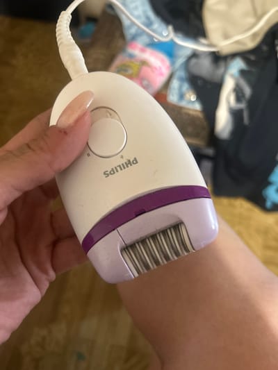 The image shows a close-up of a Philips epilator being held by a person's hand. The epilator is white with a purple trim and has a head with multiple metal tweezers. The person's thumb is resting on a circular button on the epilator. The background is blurred, but appears to be an indoor setting with some fabric items. There is no indication of the city of Perushtitsa, Bulgaria, or any specific urban environment.