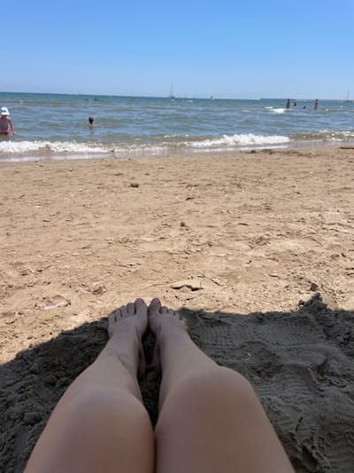 A person's legs and feet are visible on a sandy beach, with the ocean and a clear blue sky in the background. The sand is dark and appears wet in some areas, with footprints visible. The person's skin is tanned, and there are shadows cast on the sand from what is presumably the photographer. The ocean is calm with small waves rolling in. Several people are visible in the water, wading or swimming. The sun appears to be high in the sky, indicating it is daytime, likely mid-morning or early afternoon. The scene suggests a relaxing day at the beach in Valencia, Spain. There is no visible text in the image.