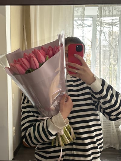 A single individual is depicted in a mirror self-portrait, captured in an indoor residential setting. While the prompt specifies Slavyansk-na-Kubani, Russia, no discernible features within the image allow for precise geographical identification of the city or a specific location within it.
The subject, partially obscured, is shown from the upper torso to just below the waist. The individual wears a black and white horizontally striped long-sleeved sweater with ribbed white cuffs. Dark hair is visible at the top of the head, and one eye and eyebrow are visible above a red-cased smartphone held in the right hand. A ring is present on the ring finger of the right hand. The individual holds a large bouquet of approximately 15-20 closed pink tulips in the left arm, wrapped in a combination of translucent clear film and light purple paper, tied with a pale pink ribbon around the stems.
The immediate background, reflected in the mirror, consists of a light-colored wall on the left, and a large window with white framing and sheer, light-colored lace curtains on the right. Beyond the window, the outdoor environment shows bare tree branches, indicative of a colder season. A dark wooden frame, likely belonging to the mirror, is visible at the top and left edges of the composition. A white electrical outlet or switch plate is partially visible on the wall to the left of the individual. The individual's action is taking a photograph of their reflection while holding the bouquet and smartphone.