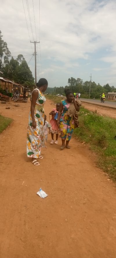 The scene takes place on a dirt road in Kivaywa, Kenya, located at latitude: 0.56, longitude: 34.806. Four people are present. Two women are walking, one wearing a white and yellow floral dress, the other wearing a blue and yellow dress with a brown bag. Two young children are walking behind the women. The children are both female, one wearing a pink dress, the other wearing a white shirt.  The children are looking at the women.  The women are facing forward, walking in the same direction. There are trees on the left side of the image, and a paved road on the right side of the image.  The sky is cloudy. There is a utility pole in the background. A piece of white trash is visible on the ground.  There are other people visible in the distance on the paved road.  A motorcycle is visible in the distance on the paved road.  The image appears to be taken from the perspective of someone walking behind the group of people.  The image appears to be taken with a mobile phone, as the resolution is low.  The quality of the image is poor.
