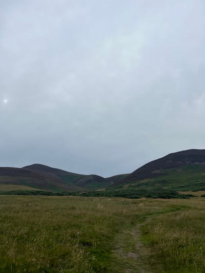 A dirt path winds through a grassy field towards rolling hills under a cloudy sky. The hills are covered in dark heather and patches of green vegetation. A faint sun can be seen in the upper left corner of the sky. The scene appears to be located in the Pentland Hills Regional Park, near Penicuik, United Kingdom. There are no people or animals visible in the image.