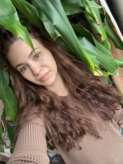 A woman with long, wavy brown hair is lying among green leaves. She is wearing a tan, ribbed sweater. Her face is tilted towards the camera, and she is smiling slightly. Her eyes are green. The background contains more green leaves and what appears to be a cardboard box. The setting appears to be indoors.
It is not possible to confirm that this image was taken in Nizhnyaya Gar', Russia. There is no information in the image to confirm the location.