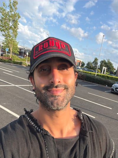 The media file shows a close-up, selfie-style shot of a person wearing a black baseball cap and a black t-shirt. The cap has a red embroidered logo with white text. The person has dark hair and a beard, and their eyes are visible. The image is taken outdoors in what appears to be a parking lot with white lines on the asphalt. In the background, there are trees, a building, and some parked cars, suggesting an urban or suburban environment. The sky is blue with scattered white clouds, indicating daytime with fair weather. The overall tone of the image is casual. Visible text on the cap reads "BOUG LE RESTAURANT & APRES-SKI."