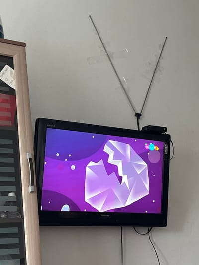 A Toshiba Regza television is mounted on a lightly textured wall. The TV screen displays a vibrant purple background with a large, abstract, crystalline-like object in shades of white and light purple. Several small white dots, resembling stars, are scattered across the screen, along with a few small, translucent circles. In the upper left corner of the screen, there are a few colorful, spherical icons.
Above and to the left of the television, a silver, V-shaped antenna is attached to the wall, with its two prongs extending outwards. A small black device is attached to the top right of the television, possibly a streaming device or a media player. Black cables are visible behind the TV, running down the wall.
Below the television, a light-colored wooden cabinet is visible, with a glass-fronted section showing vertical dark lines. A corner of a paper with text in Russian, possibly "ПРИВЕТ МИР!", and a heart symbol is visible on the cabinet. The overall scene suggests an indoor setting, likely a living room or entertainment area, given the presence of the television and cabinet. The lighting appears to be ambient, indicating daytime, but no specific weather conditions are discernible.