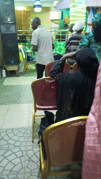 Inside a room with tile flooring, a man in a light shirt walks near a speaker. Several individuals are seated. One wears a white head covering, while another is adorned in green. The person in the foreground dons a black garment and head covering. Rows of chairs with gold frames are visible. Decorations include a chandelier and green streamers. A screen displays a colorful image in the background.
