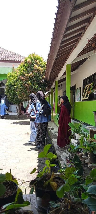 The image shows a group of four young women wearing headscarves standing in a schoolyard in Cangkuang, Indonesia (latitude: -7.065, longitude: 107.552). The women are wearing blue and red clothing. Two of the women are facing the camera, while the other two are facing away. The women are standing in front of a green building with a white roof. There are a few small trees in the foreground, and the ground is made of concrete. The scene is shot from a low angle, and the women are standing in the shade of the building. The women appear to be students and the building appears to be a school building. There are pots with plants in the foreground. The background is partly obscured by a large tree. The sky is clear and bright, with the sun shining from the top left of the image. The time appears to be daytime.