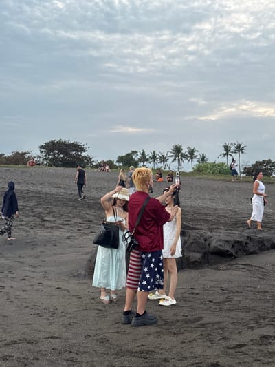 A medium shot captures a group of people on a dark sand beach with choppy, overcast skies. In the foreground, a person with bright orange hair, wearing a maroon shirt and American flag-patterned shorts, holds up a phone to record another person in a white dress. To their left, a woman in a pale blue dress and a straw hat looks at her phone while holding a black handbag. To the far right, a woman in a white top and white cropped pants walks away from the camera. In the midground, several individuals are scattered across the dark sand. Some are seated on a grassy incline dotted with trees, while others stand near palm trees in the background. One person in a dark hoodie and patterned pants walks towards the left of the frame, their back to the camera. The overall scene appears to be a public beach, possibly in the city of Mengwi, Indonesia, given the presence of palm trees and the tropical feel. The dark sand suggests a volcanic beach.
