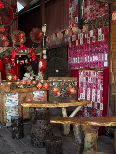 This is an indoor shot of a tourist attraction in George Town, Malaysia, likely located at the "Chew Jetty". The image is filled with decorations and objects, with an eclectic, almost chaotic arrangement.
The ceiling is adorned with traditional red and white Chinese-charactered lucky charm cards and woven baskets. There's a vintage-style electric fan mounted on the wall. The walls display traditional elements, including wooden panels with written Chinese characters, decorative plates, and red lanterns. Slogans like "RESPECT, BE YOURSELF" and "LIFE ISN'T ABOUT WAITING FOR THE STORM TO PASS" are written on wooden planks. There are also wooden stools or seats made from tree trunks and logs. The overall atmosphere is rustic.